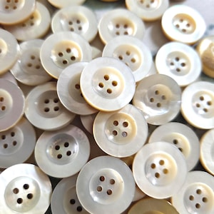 Può includere: Primo piano di una collezione di bottoni rotondi, bianco sporco. Ogni bottone ha quattro fori e un centro leggermente rialzato. I bottoni sono sparsi, creando un effetto di texture e lucentezza.