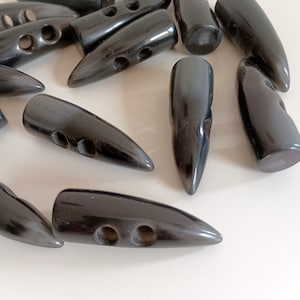 Pode incluir: Um grupo de botões de chifre preto, cada um com dois furos. Os botões têm a forma de uma bala e têm uma ponta pontiaguda.