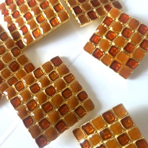 Può includere: Una collezione di bottoni quadrati caratterizzati da un motivo a griglia di tessere in smalto traslucido color ambra, arancione e marrone, incastonate in una cornice di metallo dorato. Ogni bottone presenta un design a mosaico ripetitivo con superfici lisce e lucide.