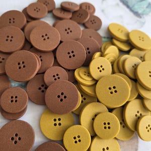 May include: A selection of round buttons in brown and mustard yellow. Each button features four small holes for sewing. The buttons are made of a matte material and are scattered across a light-coloured surface.