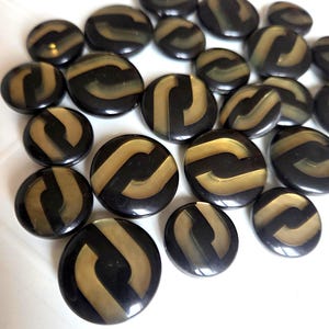 Może przedstawiać: Kolekcja czarnych i złotych guzików o unikalnym wzorze. Guziki są okrągłe, z błyszczącym wykończeniem i złotym, zakrzywionym wzorem w paski. Guziki różnią się rozmiarem i są rozrzucone na białej powierzchni.