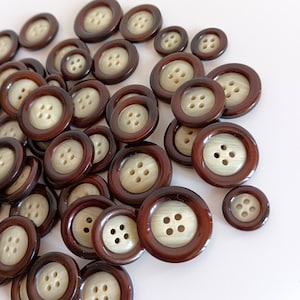 May include: A collection of brown buttons with a light beige center and four holes. The buttons vary in size and are scattered across a white surface. The buttons have a glossy finish.