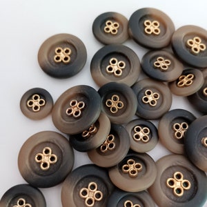 May include: A collection of brown and black buttons with gold accents. The buttons have a gradient color effect, with a darker brown color on the top and a lighter brown color on the bottom. The buttons are arranged in a random pattern on a white surface.