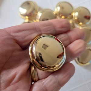 May include: A collection of shiny, gold-colored buttons. The buttons have a rounded, domed shape with a smooth, reflective surface. The buttons are approximately 1 inch in diameter. Several buttons are visible, some in focus and others blurred.