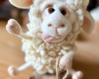 May include: A handmade, off-white sheep figurine with a playful expression. The sheep has a fluffy wool texture, pink accents on its ears and mouth, and brown eyes. It is mounted on a small wooden cart with wheels, and a string is attached.
