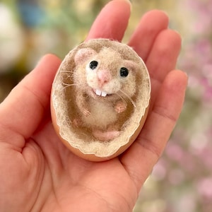 May include: A handmade felted hamster figurine inside a cracked eggshell. The hamster has a light brown body, pink nose, and black eyes. The eggshell is beige with a fluffy interior. The figurine is held in a person's hand.