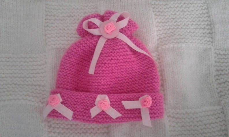 May include: Pink knitted hat with a white ribbon and pink flower embellishments.