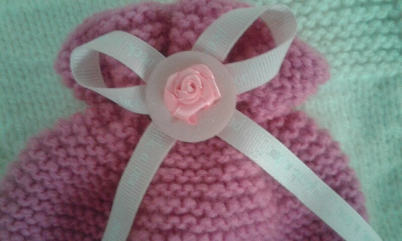 May include: A pink knitted baby hat with a white satin ribbon and a pink fabric flower.