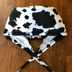 May include: A black and white cow print surgical scrub cap with ties. The cap features a white background with large black spots, and black ties for securing it. The fabric is a cotton blend.