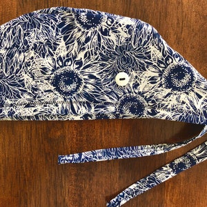 May include: A navy blue and white patterned scrub cap featuring a sunflower design. The cap has a button closure and tie straps for a secure fit. The fabric is a floral print with white outlines on a navy blue background.