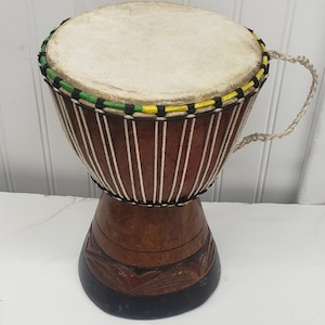 May include: A brown djembe drum with a light tan drumhead. The drum has a dark brown base with carved details and a black bottom. The top rim is decorated with green, yellow, and black accents, and a rope handle is attached.