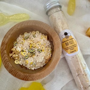 May include: A small glass jar filled with white powder and small yellow and gold decorations. The jar has a silver lid and a label that reads "Sunshine Pie Creations BEE FIZZ Pour fizz into a bowl & small amounts of water or vinegar. Ages 3+"