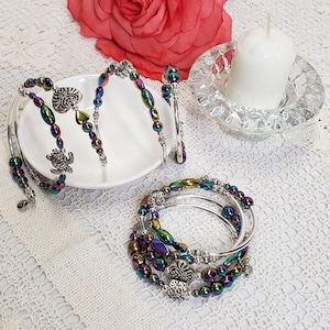 May include: A set of five iridescent bracelets with silver charms, including a butterfly, a heart, and a turtle. The bracelets are arranged on a white plate with a white background.