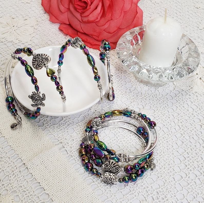 May include: A set of five iridescent bracelets with silver charms, including a butterfly, a heart, and a turtle. The bracelets are arranged on a white plate with a white background.