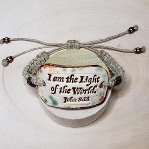 May include: A light brown hemp bracelet with a white ceramic charm that says "I am the Light of the World, John 8:12".