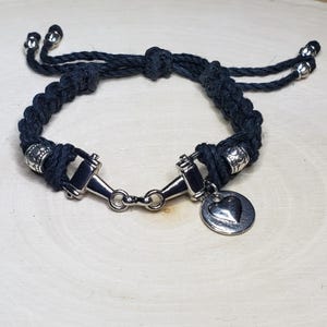 May include: Black braided rope bracelet with silver-toned metal accents. The bracelet features a horse bit design and a small heart charm. Adjustable sliding knot closure with silver beads on the ends.