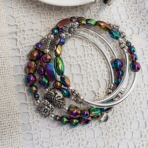 May include: A set of four iridescent silver bangles with decorative charms. The charms include a butterfly, a flower, and a heart. The bangles are stacked on top of each other and are arranged in a circular pattern.