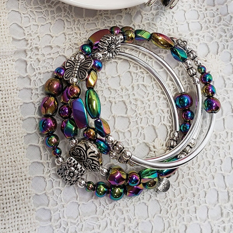 May include: A set of four iridescent silver bangles with decorative charms. The charms include a butterfly, a flower, and a heart. The bangles are stacked on top of each other and are arranged in a circular pattern.