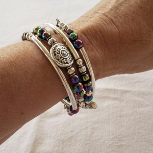 May include: A set of four silver bracelets with iridescent beads and decorative charms. The bracelets are layered on a person's wrist.