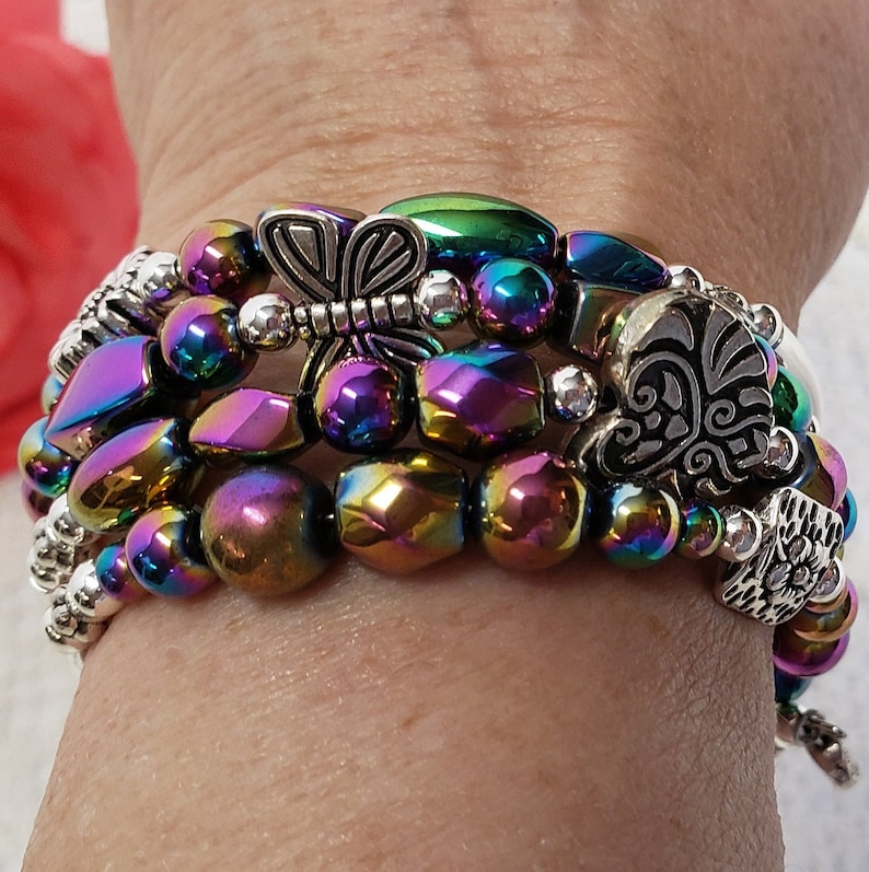 May include: A multi-strand bracelet featuring iridescent beads and silver charms. The charms include a dragonfly, a heart, and a stylised wing. The bracelet is made of multiple strands of beads, each with a different colour.