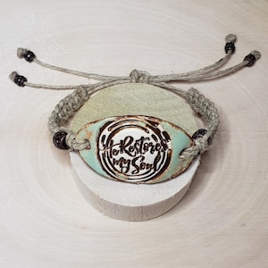 May include: A handmade bracelet with a woven cord and an oval, turquoise-colored centerpiece. The centerpiece features the phrase "She Restores My Soul" in a dark, stylized font. The bracelet is displayed on a light green stone.