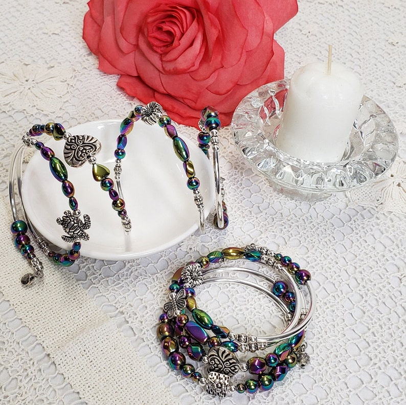 May include: A set of five iridescent beaded bracelets with silver charms. The bracelets are stacked on top of each other and feature a variety of charms, including a heart, a turtle, and a flower.