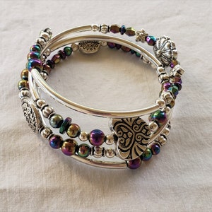 May include: A silver bangle bracelet with iridescent beads and silver charms. The bracelet is made of three strands of beads and has a heart-shaped charm, a flower charm, and a round charm.