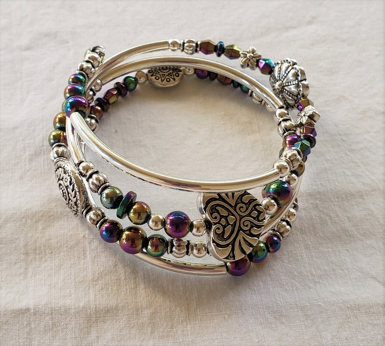 May include: A silver bangle bracelet with iridescent beads and silver charms. The bracelet is made of three strands of beads and has a heart-shaped charm, a flower charm, and a round charm.