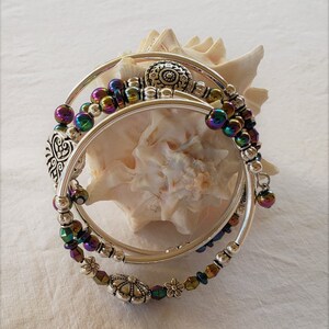 May include: Three silver bangles with iridescent beads and decorative charms. The bangles are stacked on top of each other and are resting on a seashell.