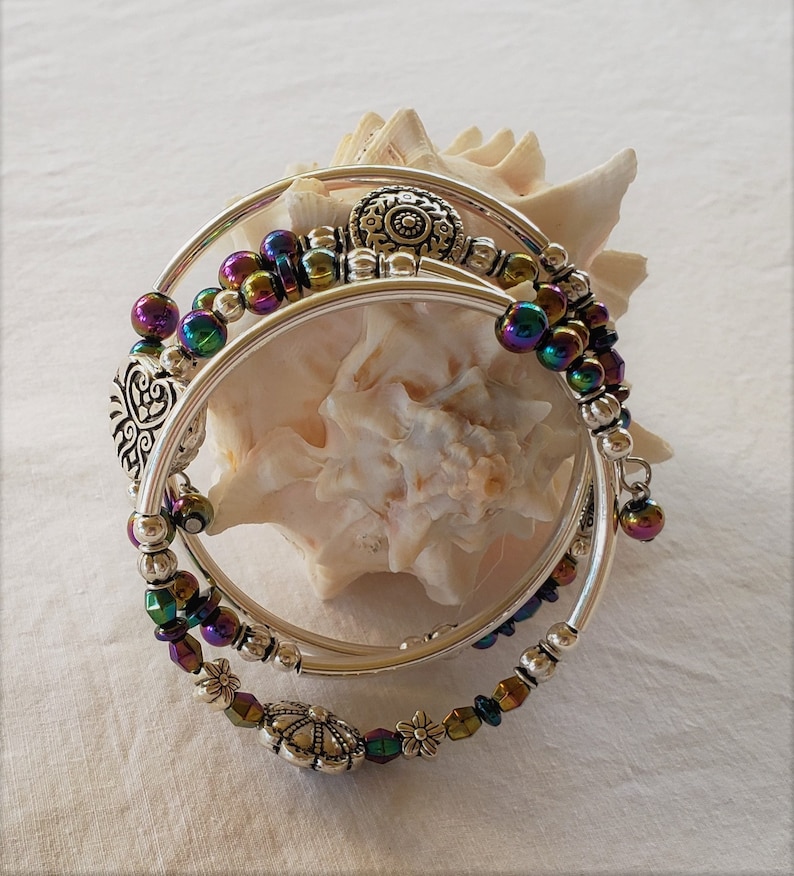 May include: Three silver bangles with iridescent beads and decorative charms. The bangles are stacked on top of each other and are resting on a seashell.