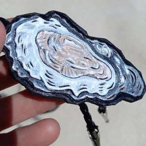 May include: A black leather bolo tie with a white and silver painted oyster shell design.