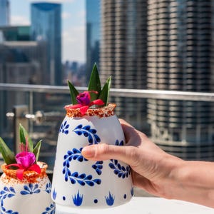 May include: A white ceramic cocktail glass with blue floral designs, held by a hand. The rim is coated with a red spice. Garnished with green leaves, a pink flower, and a cherry. The background features a cityscape.