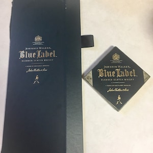 May include: A dark blue box with a gold Johnnie Walker Blue Label blended scotch whisky logo. The box is closed and has a black ribbon attached to the top. A small, square, dark blue and gold Johnnie Walker Blue Label blended scotch whisky coaster is also shown.