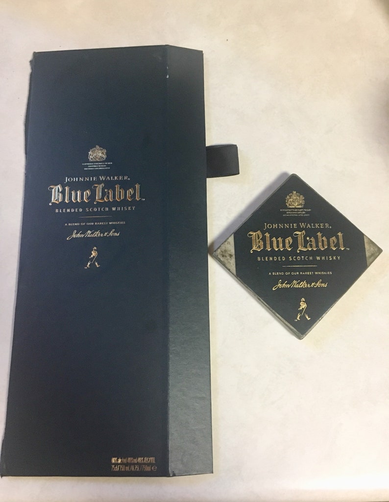 May include: A dark blue box with a gold Johnnie Walker Blue Label blended scotch whisky logo. The box is closed and has a black ribbon attached to the top. A small, square, dark blue and gold Johnnie Walker Blue Label blended scotch whisky coaster is also shown.