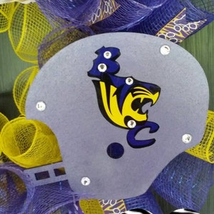 Puede incluir: Una corona de malla morada y amarilla con un casco de f&uacute;tbol gris con una mascota amarilla y azul. El casco est&aacute; decorado con diamantes de imitaci&oacute;n. Un coraz&oacute;n de cebra en blanco y negro con el texto "gurl gone craft" est&aacute; unido a la corona.