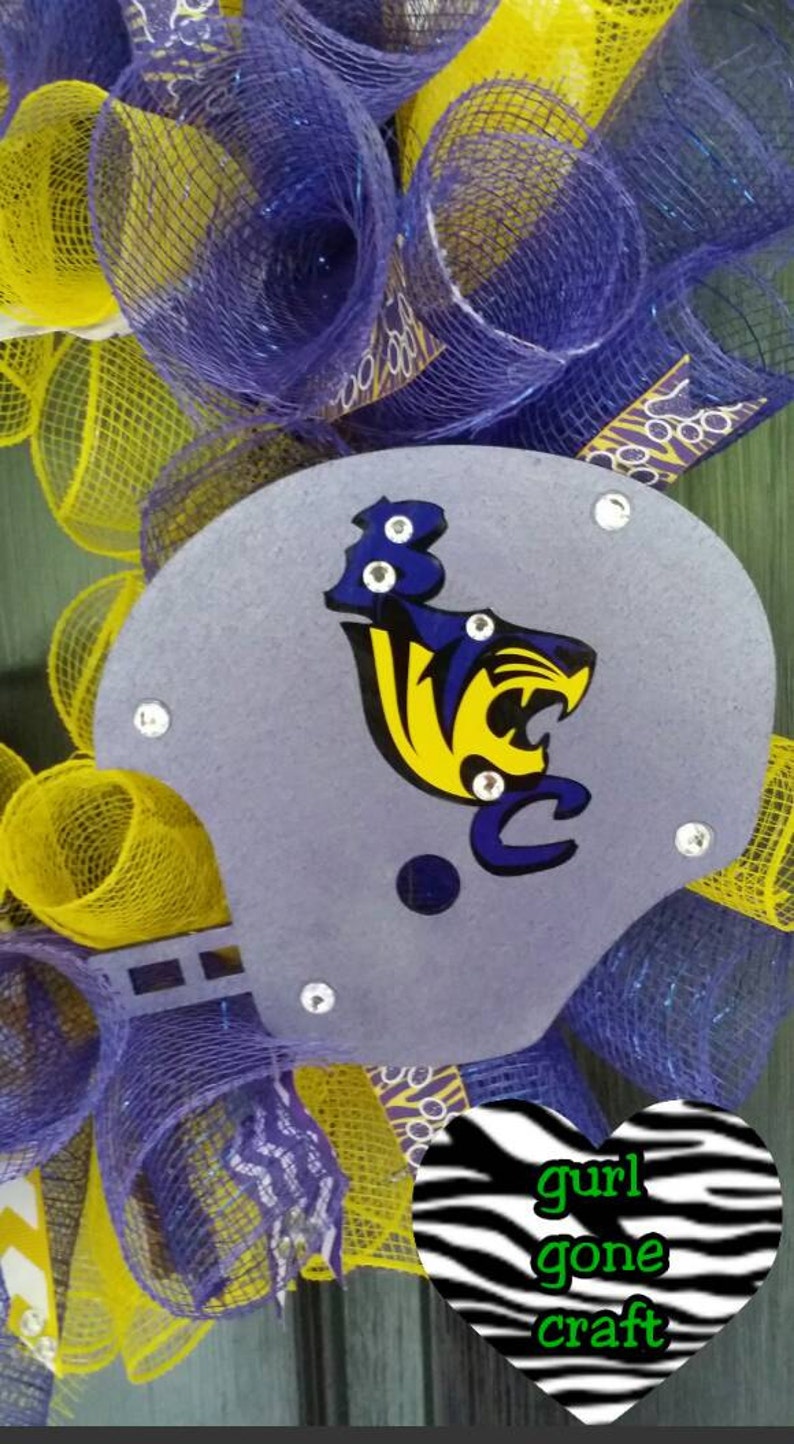 Puede incluir: Una corona de malla morada y amarilla con un casco de f&uacute;tbol gris con una mascota amarilla y azul. El casco est&aacute; decorado con diamantes de imitaci&oacute;n. Un coraz&oacute;n de cebra en blanco y negro con el texto "gurl gone craft" est&aacute; unido a la corona.