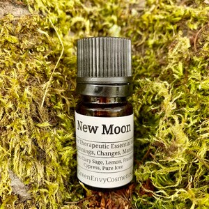 May include: A small brown glass bottle with a black cap, labelled "New Moon Therapeutic Essential Oil for Beginnings, Changes, Manifestations. Clary Sage, Lemon, Frankincense, Cypress, Pure Love. Green Envy Cosmetics."