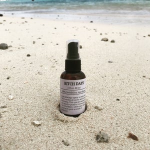 Small brown bottle with a white label and a black spray top, nestled in the sand on a beach with rocks in the sand and the ocean in the background.