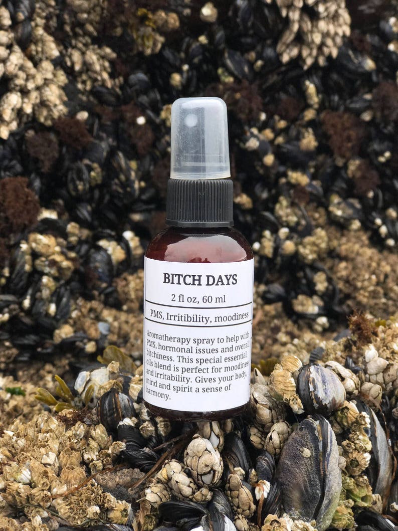 May include: A brown glass bottle with a black spray top and white label. The label reads "BITCH DAYS 2 fl oz, 60 ml PMS, Irritability, moodiness Aromatherapy spray to help with PMS, hormonal issues and overall bitchiness. This special essential oil blend is perfect for moodiness and irritability. Gives your body, mind and spirit a sense of harmony."