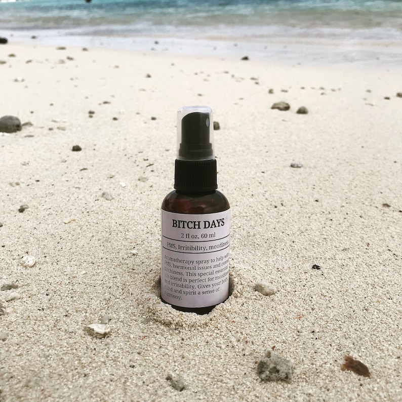 Small brown bottle with a white label and a black spray top, nestled in the sand on a beach with rocks in the sand and the ocean in the background.