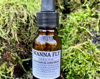 Puede incluir: Una botella de vidrio marrón de aceite de viaje astral "WANNA FLY" con un cuentagotas negro. La etiqueta indica que el aceite está infundido con cristales, 15 ml. La botella está colocada sobre un fondo de musgo verde.