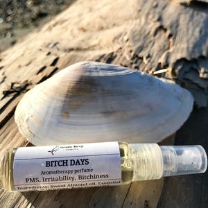 May include: A small clear glass bottle with a white label that reads "BITCH DAYS Aromatherapy perfume PMS, Irritability, Bitchiness Ingredients: Sweet Almond oil, Essential"