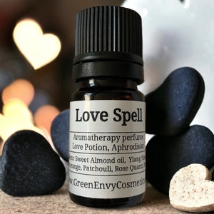 May include: A small glass bottle with a black cap and a white label that reads "Love Spell Aromatherapy perfume Love Potion, Aphrodisiac ats: Sweet Almond oil, Ylang trange, Patchouli, Rose Quartz, W.Green Envy Cosmetics". The bottle is surrounded by black and white heart-shaped stones.
