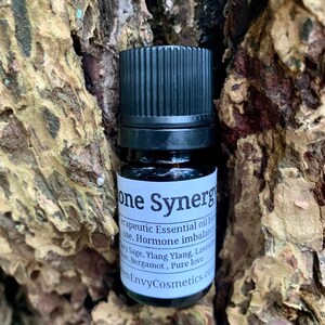 May include: A small brown glass bottle with a black cap and a white label that reads "one Synergy therapeutic Essential oil blend use, Hormone imbalance Clary Sage, Ylang Ylang, Lavender, Bergamot, Pure love en EnvyCosmetics.com".