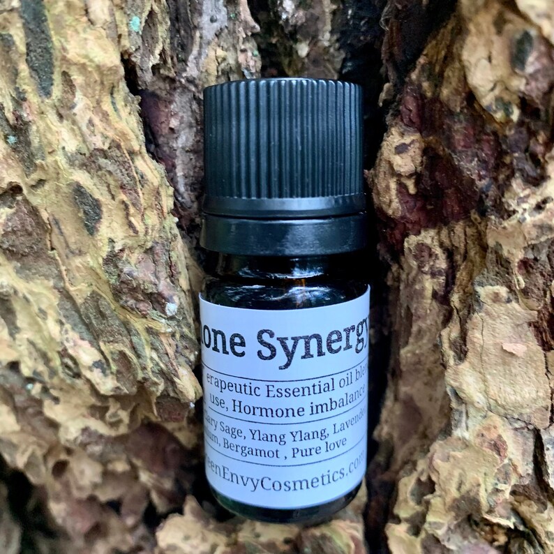 May include: A small brown glass bottle with a black cap and a white label that reads "one Synergy therapeutic Essential oil blend use, Hormone imbalance Clary Sage, Ylang Ylang, Lavender, Bergamot, Pure love en EnvyCosmetics.com".