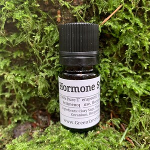 May include: A small brown glass bottle with a black cap, labeled "Hormones 100% Pure Therapeutic Essential Oils Perimenol use, Hormone Balance" and "Ingredients: Clary Sage, Ylang Ylang, Geranium, Bergamot" with the website address "www.GreenEnvyCo.com".