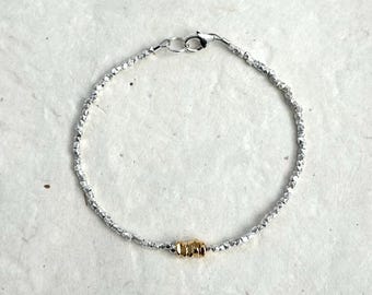 Puede incluir: Una pulsera delicada compuesta por pequeñas cuentas plateadas facetadas dispuestas en una cadena circular. La pulsera presenta un grupo de cuentas doradas como acento en la parte inferior y un cierre de mosquetón plateado.