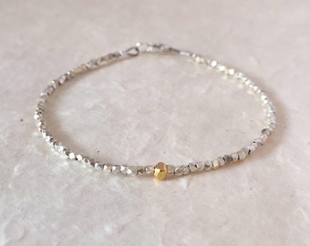 Puede incluir: Un delicado brazalete de plata con un solo acento de cuentas doradas. El brazalete está hecho de pequeñas cuentas facetadas y tiene un diseño simple y elegante.