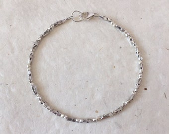Puede incluir: Una delicada pulsera de plata con pequeñas cuentas facetadas ensartadas. La pulsera tiene un diseño sencillo con cierre de mosquetón. Las cuentas tienen una superficie texturizada y reflectante, creando un brillo sutil. La pulsera mide aproximadamente 18 cm de largo.