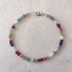 May include: A multi-colored gemstone bracelet with a silver clasp. The bracelet features beads of amethyst, aquamarine, carnelian, emerald, ruby, and white pearls.